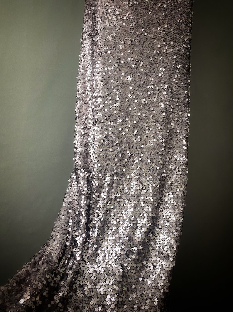 May include: A silver sequined fabric with a subtle sheen. The fabric is draped and shows the texture of the sequins.