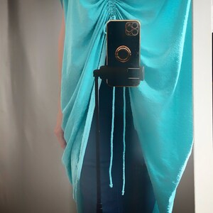 May include: A turquoise blue, loose-fitting, long tunic top with a drawstring at the neckline. The top is draped over a pair of dark blue jeans.