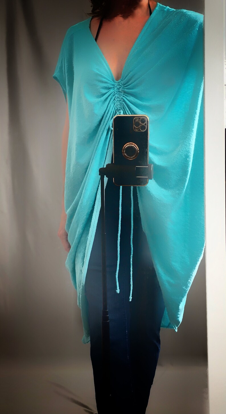 May include: A turquoise blue, loose-fitting, long tunic top with a drawstring at the neckline. The top is draped over a pair of dark blue jeans.