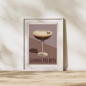 Peut inclure: Impression d'art encadrée représentant un cocktail dans un verre à pied. La boisson a une mousse sur le dessus et est garnie de grains de café. Le fond est un dégradé de marron et de violet, avec le texte "GAMMA PHI BETA" en bas.