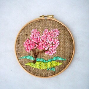 May include: A round embroidery hoop featuring a hand-stitched tree with pink blossoms, a brown trunk, and green grass. The artwork is set against a burlap background and framed by a wooden hoop.