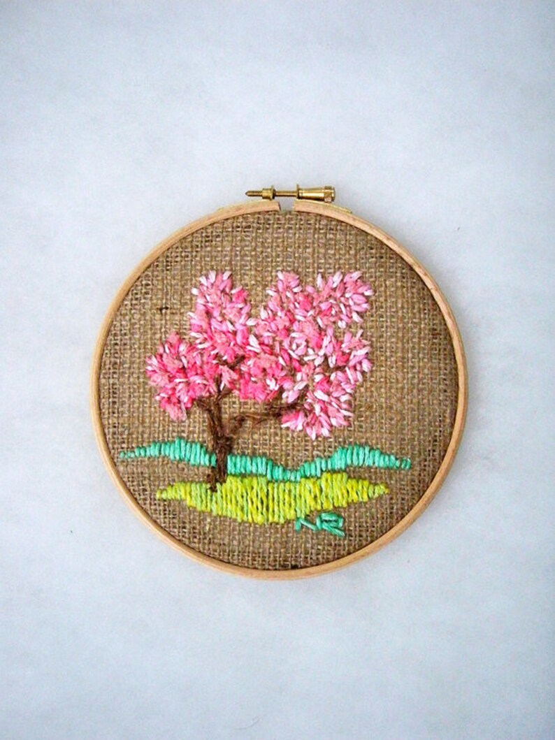 May include: A round embroidery hoop featuring a hand-stitched tree with pink blossoms, a brown trunk, and green grass. The artwork is set against a burlap background and framed by a wooden hoop.
