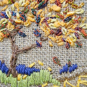 May include: Close-up of an embroidered tree scene on a burlap-like fabric. The tree has a brown trunk and colorful leaves in shades of yellow, orange, and green. The ground features blue and green embroidered elements, with a gold border.