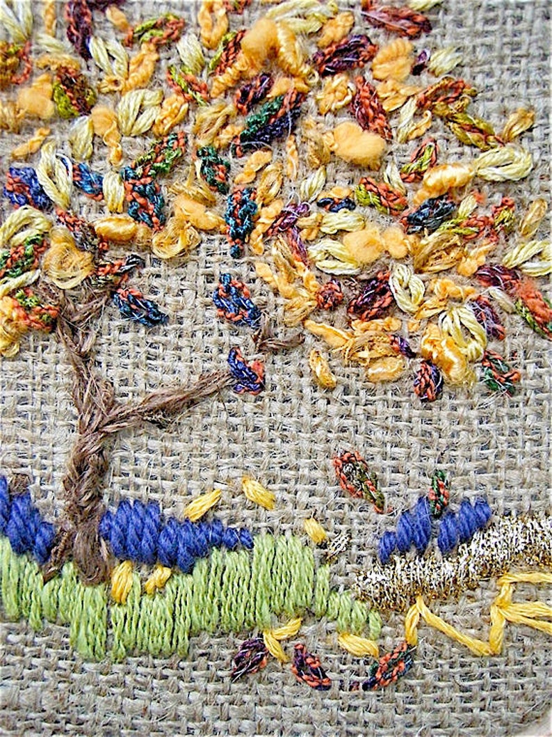 May include: Close-up of an embroidered tree scene on a burlap-like fabric. The tree has a brown trunk and colorful leaves in shades of yellow, orange, and green. The ground features blue and green embroidered elements, with a gold border.