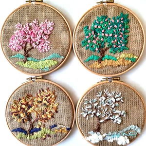 May include: Four embroidered tree art pieces in wooden hoops. Each features a different season: spring with pink blossoms, summer with green leaves, autumn with yellow and brown foliage, and winter with white snow. The background is a burlap fabric.