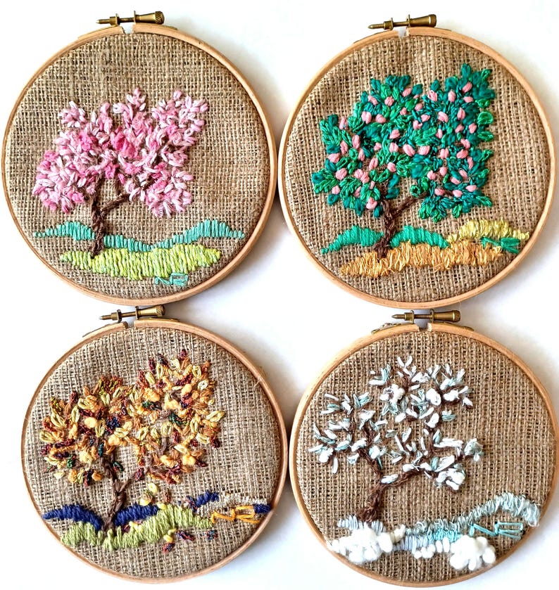 May include: Four embroidered tree art pieces in wooden hoops. Each features a different season: spring with pink blossoms, summer with green leaves, autumn with yellow and brown foliage, and winter with white snow. The background is a burlap fabric.