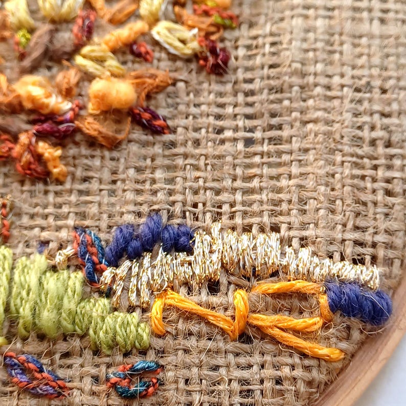 May include: Close-up of an embroidered artwork on burlap fabric. The embroidery features a variety of colors, including orange, blue, green, and gold. The stitches create a textured, detailed design, showcasing the craft of hand embroidery.