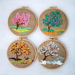 May include: Four round embroidery hoops each featuring a different tree design. The trees are embroidered with colorful thread on a burlap background. The trees depict different seasons: spring, summer, autumn, and winter. Each hoop is framed with a wooden ring.