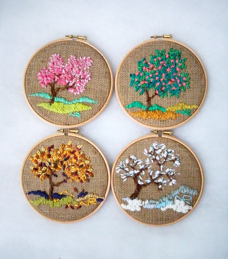 May include: Four round embroidery hoops each featuring a different tree design. The trees are embroidered with colorful thread on a burlap background. The trees depict different seasons: spring, summer, autumn, and winter. Each hoop is framed with a wooden ring.