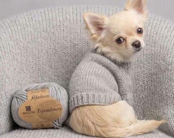 May include: A small, light-colored dog with large ears wearing a light gray knitted sweater. Beside the dog sits a ball of light gray yarn with a paper label that reads "bfashion" and "Ecocashmere" in dark brown text.