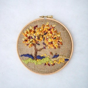 May include: A handcrafted embroidery of a tree in a wooden hoop. The tree features a brown trunk and branches with colorful yellow, orange, and red leaves. The base of the tree has green and blue stitched details, with falling leaves.