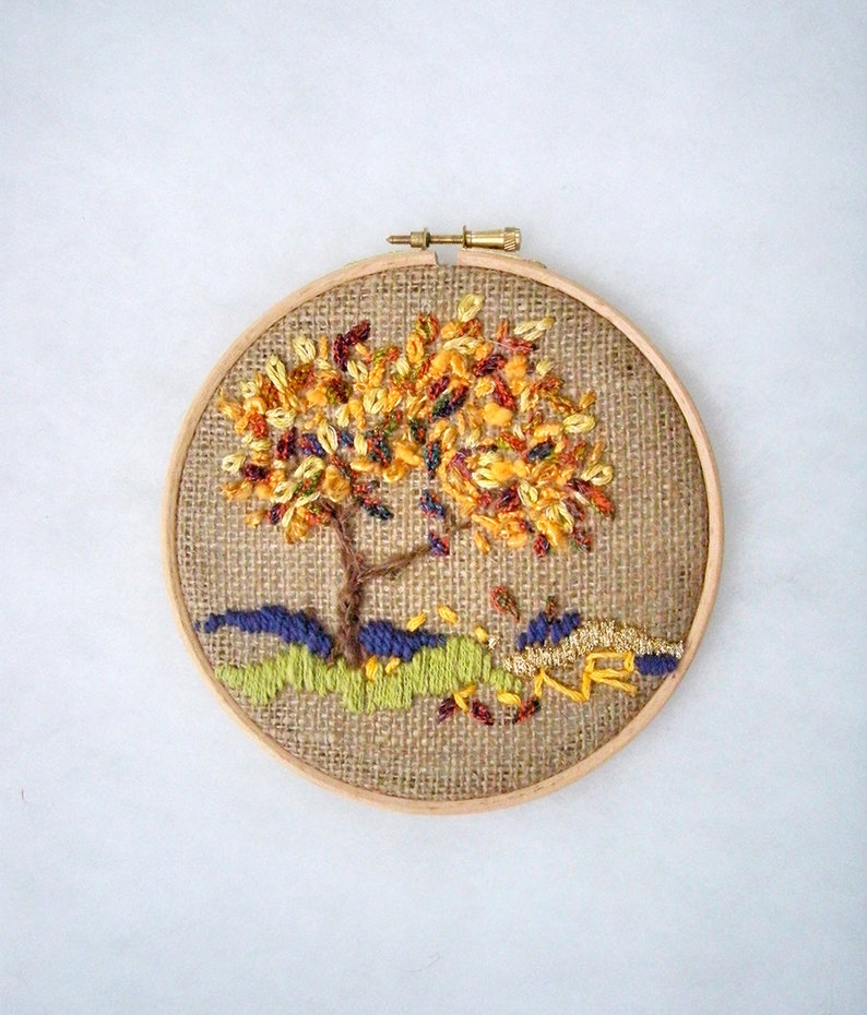 May include: A handcrafted embroidery of a tree in a wooden hoop. The tree features a brown trunk and branches with colorful yellow, orange, and red leaves. The base of the tree has green and blue stitched details, with falling leaves.