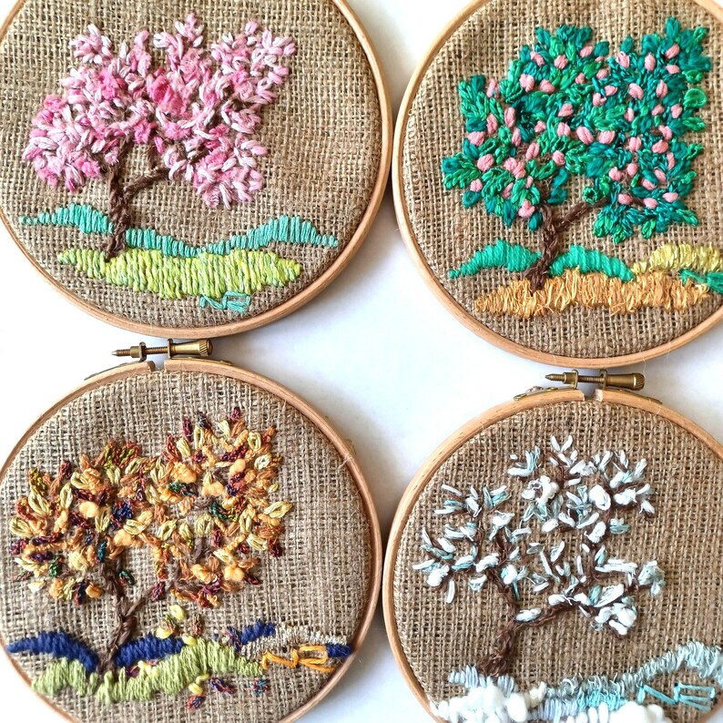 May include: Four embroidered tree designs in wooden hoops. Each features a different season: spring with pink blossoms, summer with green leaves, autumn with yellow and orange foliage, and winter with white flowers. The background is a natural burlap fabric.
