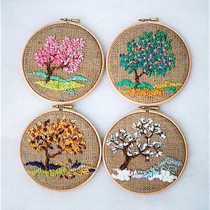 May include: Four round embroidery hoops with embroidered tree designs. Each tree represents a different season: spring with pink blossoms, summer with green leaves, autumn with golden foliage, and winter with white snow. The background is burlap.