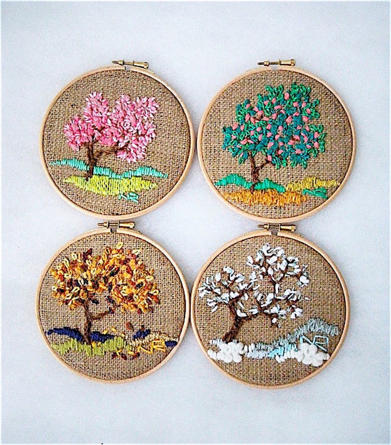 May include: Four round embroidery hoops with embroidered tree designs. Each tree represents a different season: spring with pink blossoms, summer with green leaves, autumn with golden foliage, and winter with white snow. The background is burlap.