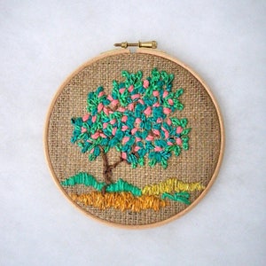 May include: Embroidered tree art in a wooden hoop. The tree features a brown trunk, teal leaves, and pink blossoms. The ground is depicted with green and yellow thread. The background is a burlap fabric.