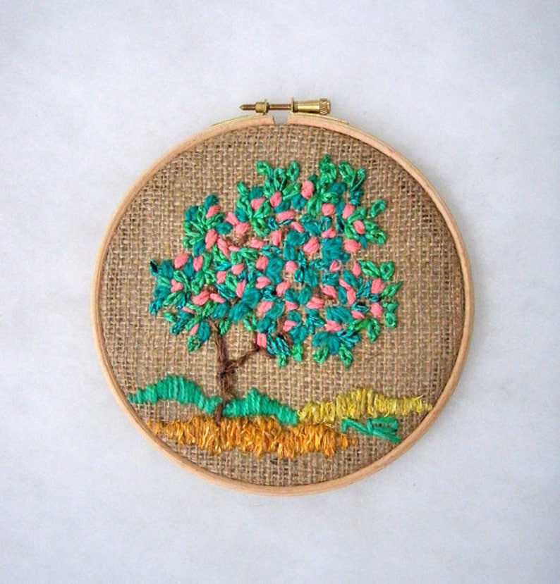 May include: Embroidered tree art in a wooden hoop. The tree features a brown trunk, teal leaves, and pink blossoms. The ground is depicted with green and yellow thread. The background is a burlap fabric.