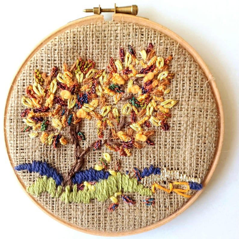 May include: An embroidered tree with autumn-colored leaves and a brown trunk, set against a burlap background. The tree is stitched within a wooden embroidery hoop. The base of the tree features blue and green embroidered details, adding depth to the design.