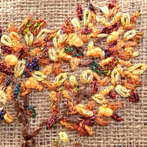 May include: Close-up of an embroidered tree with a brown trunk and branches. The leaves are a mix of yellow, orange, green, and red threads, creating a textured, autumn-themed design on a burlap background.