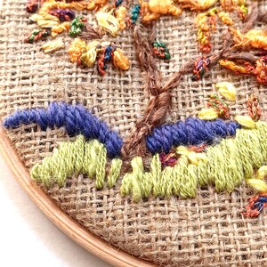 May include: Close-up of an embroidered tree on burlap fabric. The tree features a brown trunk, with yellow, orange, and green leaves. Blue and green stitched details form the ground, all set within a wooden embroidery hoop.