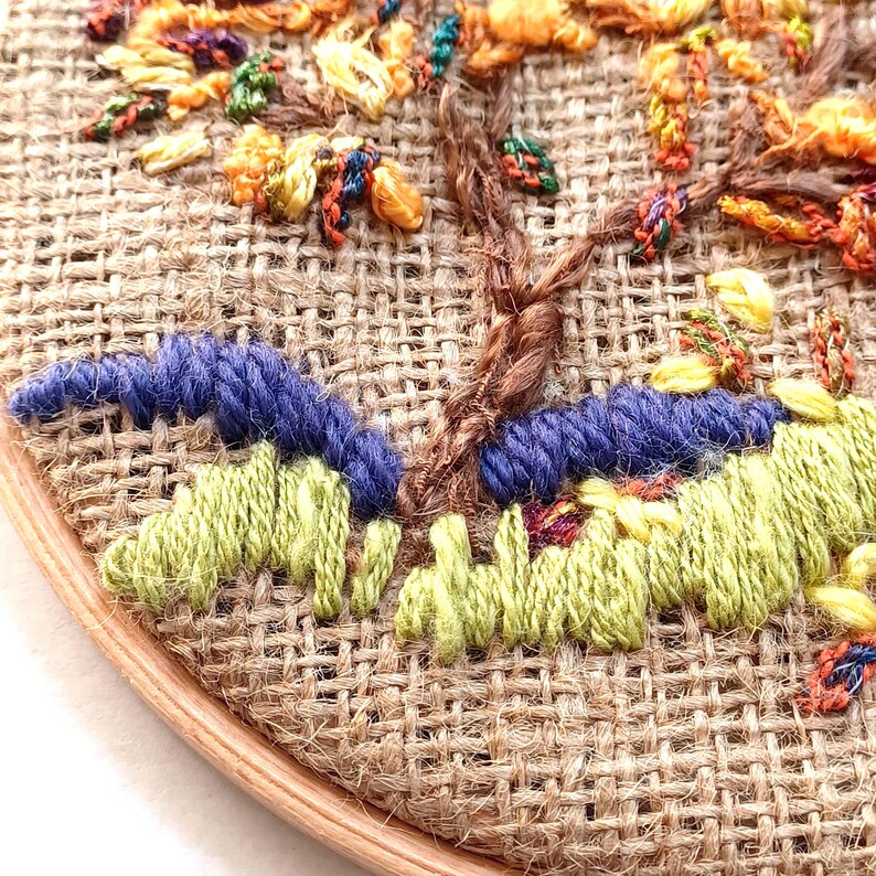 May include: Close-up of an embroidered tree on burlap fabric. The tree features a brown trunk, with yellow, orange, and green leaves. Blue and green stitched details form the ground, all set within a wooden embroidery hoop.