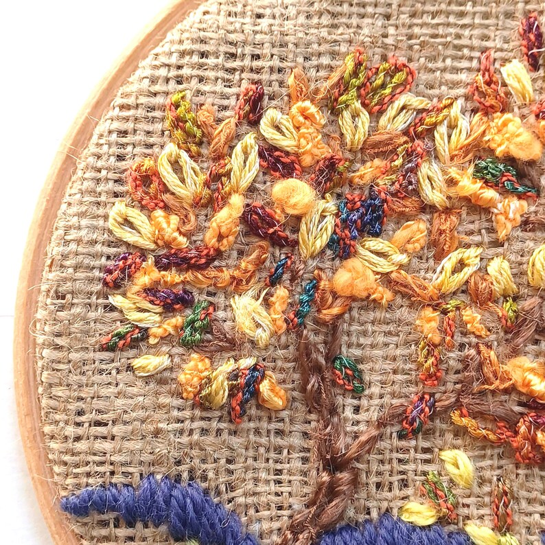 May include: Hand-stitched embroidery art of a tree with colorful leaves in shades of orange, yellow, green, and red. The tree trunk is brown, and the base is blue. The embroidery is set in a wooden hoop on a burlap background.