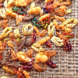 May include: Close-up of textured embroidery in shades of orange, yellow, brown, and green. The embroidery is on a burlap-like fabric, with various stitches creating a detailed, tactile surface. The colors and textures suggest a fall or harvest theme.