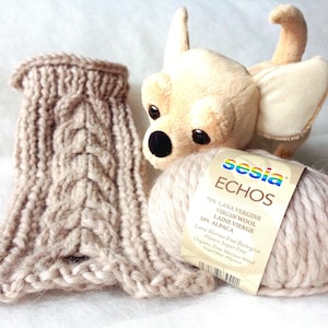 Puede incluir: Un ovillo de lana beige con una etiqueta que dice "sesia ECHOS 70% LANA VERGINE VIRGIN WOOL 30% ALPACA LAINE VIERGE Lana Merino Fine Biologica Organic Fine Merino Wool Alpaca Super Fine Superfine Alpaca". En el fondo, una bufanda de punto beige y un perro de peluche beige.