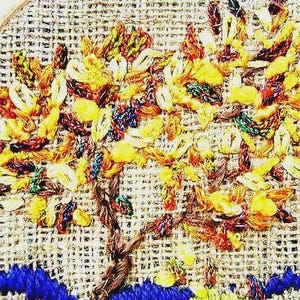 May include: Embroidered tree art with a brown trunk, yellow and orange leaves, and blue accents. The artwork is on a beige woven background, creating a textured appearance. The tree is the central focus of the piece.