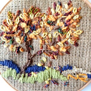 May include: Hand-stitched embroidery art of a tree with colorful leaves in shades of orange, yellow, and red. The tree trunk is brown, and the base features blue and green stitched details. The artwork is set in a round wooden frame.