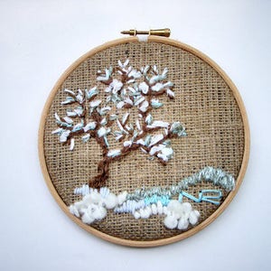 May include: Embroidered tree art in a wooden hoop. The tree has a brown trunk and branches with white and light blue accents, set against a burlap background. The artwork is a unique, handmade piece.