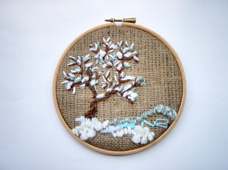 May include: Embroidered tree art in a wooden hoop. The tree has a brown trunk and branches with white and light blue accents, set against a burlap background. The artwork is a unique, handmade piece.