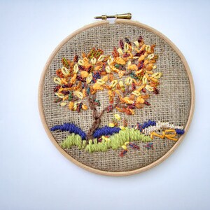 May include: Hand-stitched embroidery art in a wooden hoop. The design features a tree with colorful autumn foliage, brown trunk, and green and blue grass. The background is a burlap fabric.