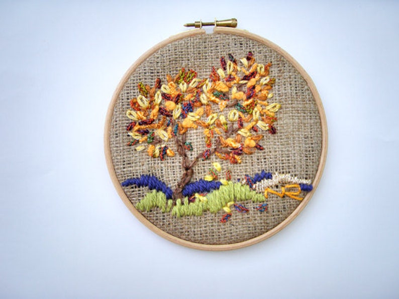 May include: Hand-stitched embroidery art in a wooden hoop. The design features a tree with colorful autumn foliage, brown trunk, and green and blue grass. The background is a burlap fabric.