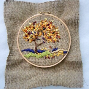 May include: An embroidered tree with colorful leaves in a wooden hoop, set against a burlap background. The tree features a brown trunk and branches, with yellow, orange, and red leaves. The base of the tree has green and blue embroidered details.