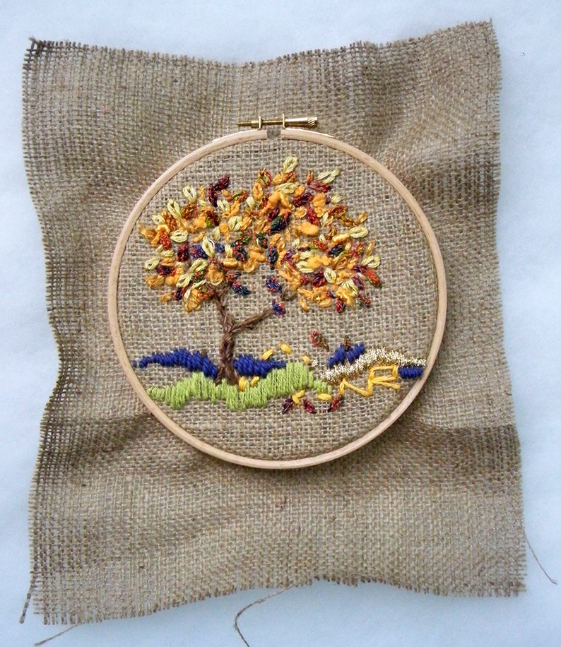 May include: An embroidered tree with colorful leaves in a wooden hoop, set against a burlap background. The tree features a brown trunk and branches, with yellow, orange, and red leaves. The base of the tree has green and blue embroidered details.
