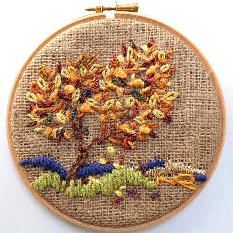 May include: Hand-stitched embroidery art featuring a tree with autumn-colored leaves. The tree is set against a burlap background within a wooden hoop. The base of the tree is surrounded by green and blue stitched details, creating a landscape scene.