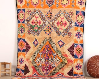 May include: A large rectangular area rug featuring a tan background with a vibrant geometric pattern in orange, pink, blue, purple, and yellow. The design includes large central diamonds and smaller repeating motifs, finished with a fringed edge.