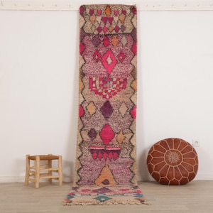 May include: A long, narrow runner rug featuring a vibrant, multi-colored geometric pattern in shades of magenta, purple, orange, and teal against a natural beige background. The rug has a textured, hand-knotted appearance with a fringed bottom edge.