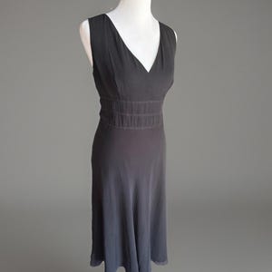 Puede incluir: Un vestido sin mangas gris oscuro con cuello en V y cintura entallada. El vestido tiene una falda fluida que llega hasta la pantorrilla. El vestido se exhibe en un maniqu&iacute; blanco sobre un fondo gris. El vestido est&aacute; hecho de una tela suave y fluida.