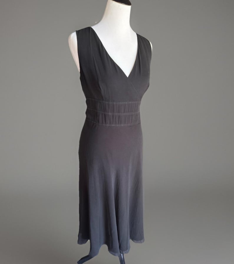 Puede incluir: Un vestido sin mangas gris oscuro con cuello en V y cintura entallada. El vestido tiene una falda fluida que llega hasta la pantorrilla. El vestido se exhibe en un maniqu&iacute; blanco sobre un fondo gris. El vestido est&aacute; hecho de una tela suave y fluida.