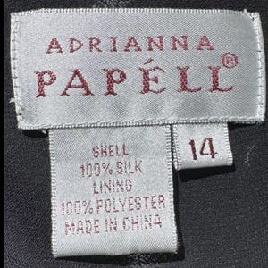 Puede incluir: Primer plano de una etiqueta de ropa con el nombre de la marca "Adrianna Papell" en letras burdeos. La etiqueta indica que la prenda es de talla 14, con una cubierta de 100% seda y un forro de 100% poli&eacute;ster. La etiqueta dice "Made in China."