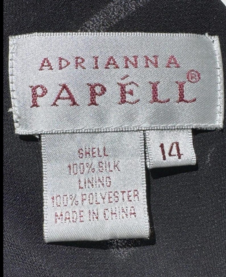Puede incluir: Primer plano de una etiqueta de ropa con el nombre de la marca "Adrianna Papell" en letras burdeos. La etiqueta indica que la prenda es de talla 14, con una cubierta de 100% seda y un forro de 100% poli&eacute;ster. La etiqueta dice "Made in China."