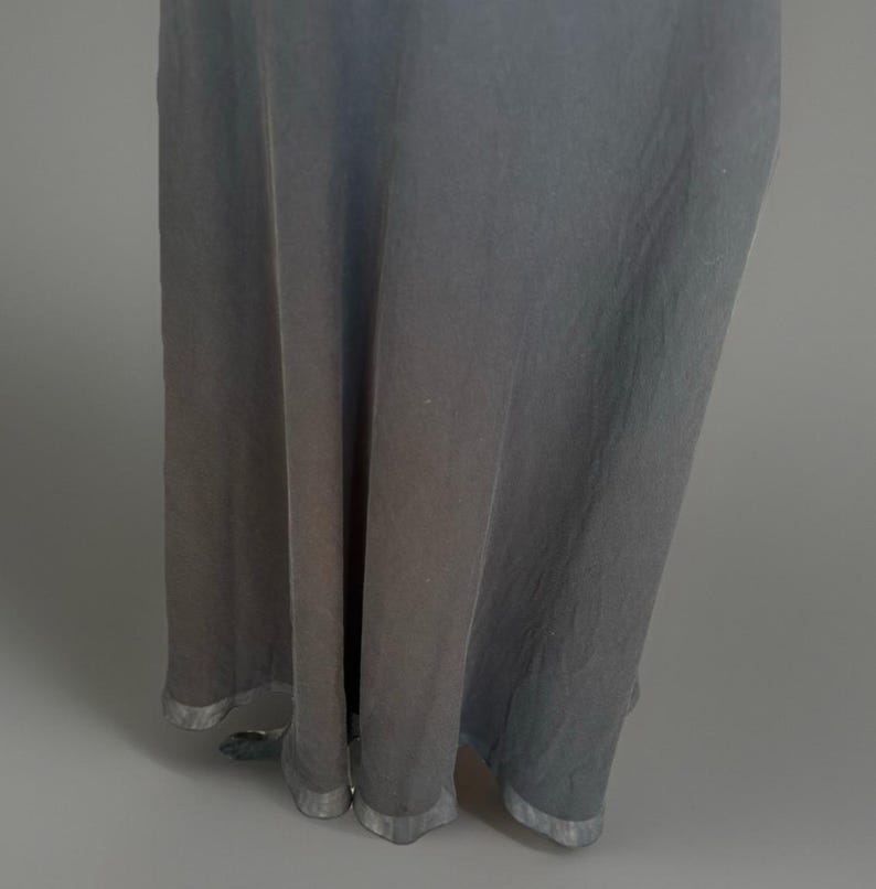Puede incluir: Un vestido largo y fluido en un tono gris apagado. La tela cae en suaves pliegues, con un brillo sutil. El dobladillo est&aacute; terminado con un ribete plateado, agregando un toque de elegancia al dise&ntilde;o simple. Ideal para eventos.