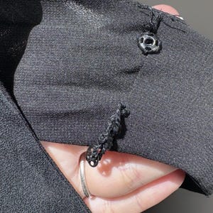 Puede incluir: Primer plano de tela negra con dos botones decorativos. Los botones son negros con un borde plateado. La tela parece ser un material suave y tejido. La imagen est&aacute; bien iluminada, destacando la textura y los detalles de la tela y los botones.