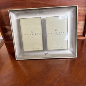 May include: A silver-toned double photo frame with two openings, each holding a cream-colored insert. The frame is labeled "Pottery Barn sentiment frame" and is designed for 4 x 6 inch photos. The word "angels" is printed at the bottom.
