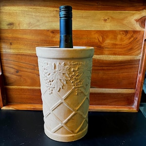 May include: A beige ceramic wine chiller with a diamond pattern and grape vine relief. A dark wine bottle is inside the chiller. The chiller is on a black surface, with a wooden tray in the background.