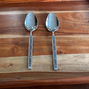 May include: Two stainless steel spoons with oval bowls and decorative handles. The handles feature an embossed pattern. The spoons are displayed on a wooden tray with a rich, warm tone.