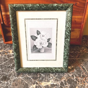 May include: A rectangular picture frame with a dark green faux marble finish containing a black and white print of a flower arrangement in a vase. The print is centered within a white mat board featuring a thin decorative border.