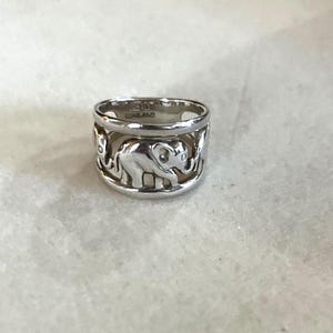 May include: A silver ring with a repeating elephant design. The ring features a band with cut-out elephant silhouettes. The ring is a wide band style and appears to be made of a shiny metal.